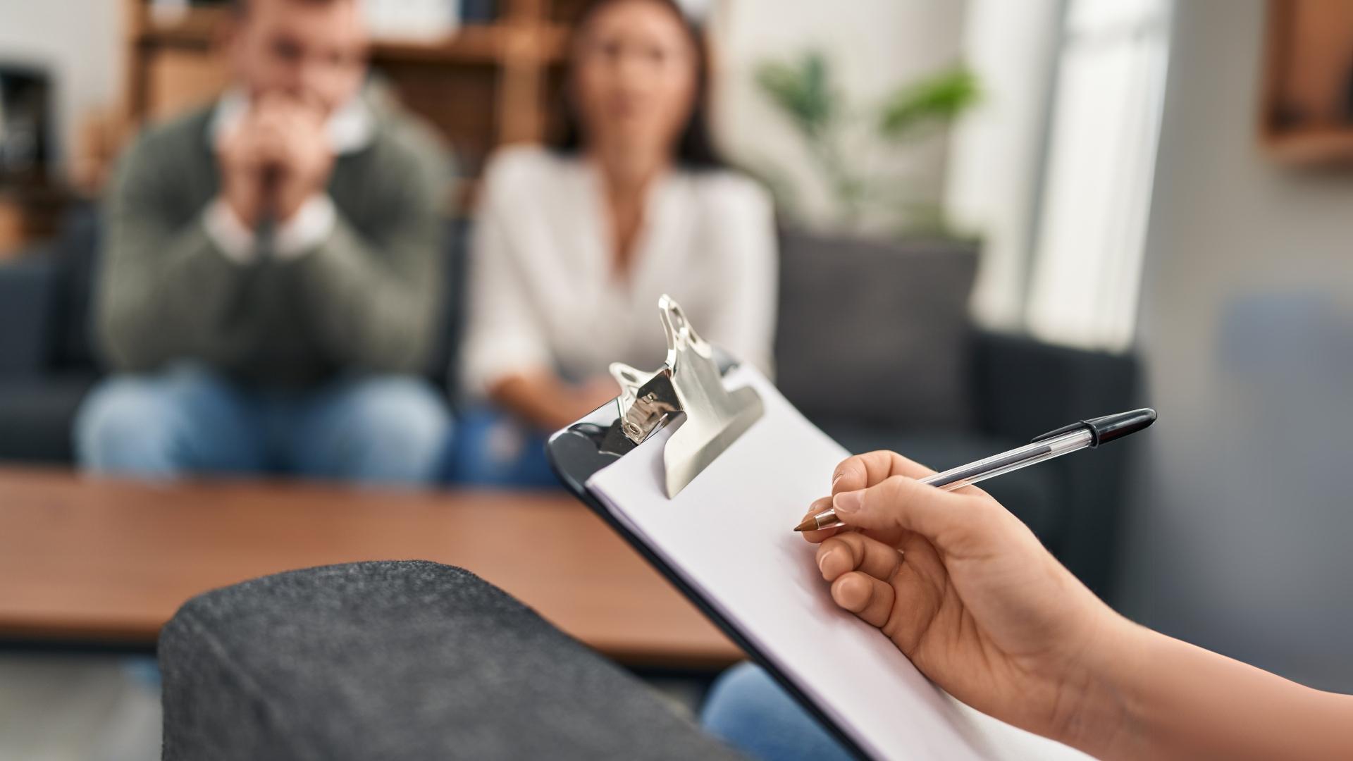 Therapist taking notes on a clipboard during a counseling session with a blurred couple in the background.