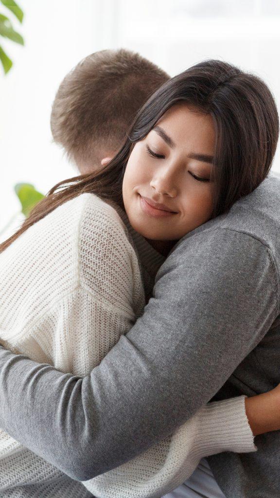 A close-up of a young woman with long dark hair, wearing a white knit sweater, warmly hugging a man in a grey sweater. She has a peaceful, gentle smile with her eyes closed, conveying comfort and emotional connection. The background is softly lit, creating a calm, intimate atmosphere.
