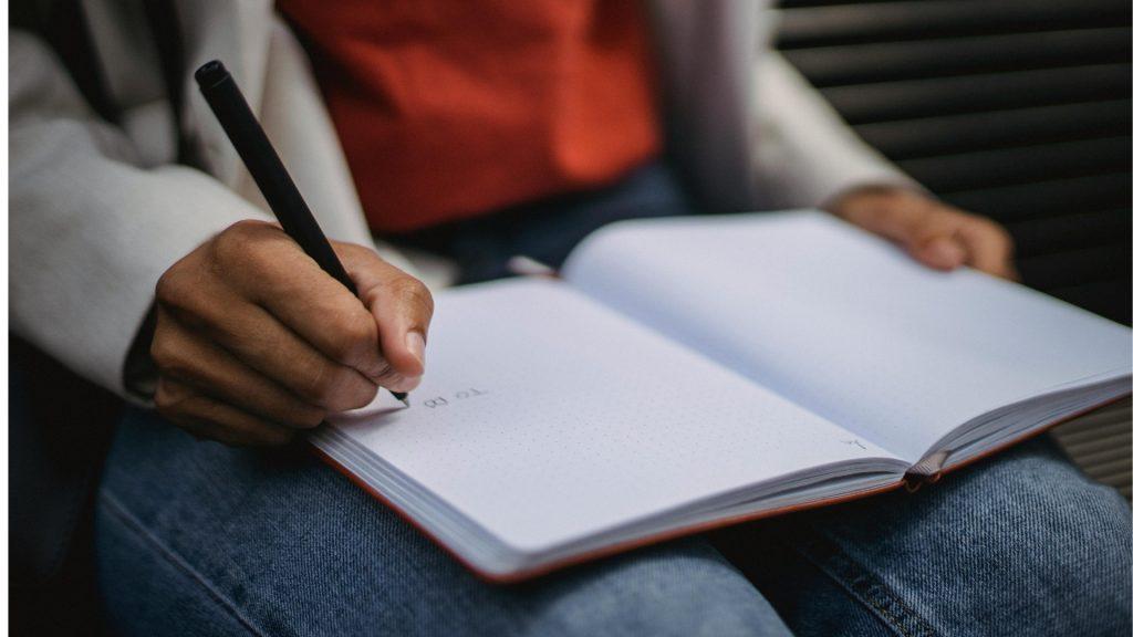 Person writing in a blank dotted notebook with a black pen.