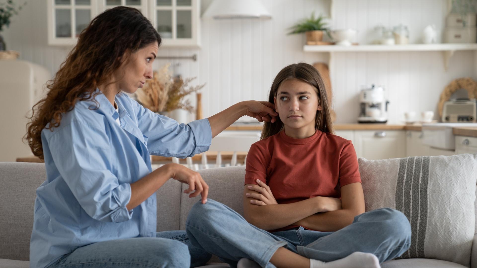 A mother and teenage daughter sit on a couch at home having a tense conversation. The teen looks upset and avoids eye contact while the mother tries to talk, reflecting the challenges parents face when supporting an anxious teen.